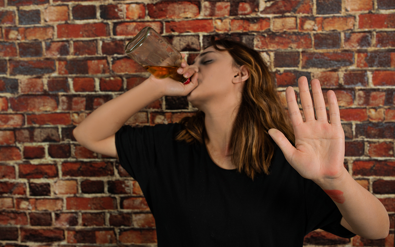 a drunk women drinking whiskey straight from the bottle in front of a red brick wall