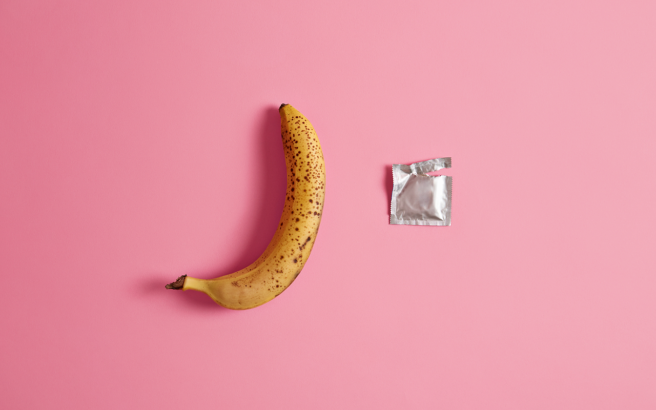 Unpacked condom and one whole banana on pink studio background. Contraceptives, prevention of venereal disease, preventing pregnancy, birth control. Reversible, temporary form of contraception.